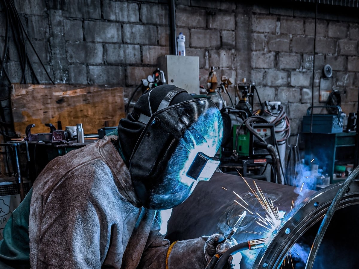 A welder in a helmet welding around a round steel section held in a fixture &mdash; representative imagery, not a specific Late Night Fabrication job