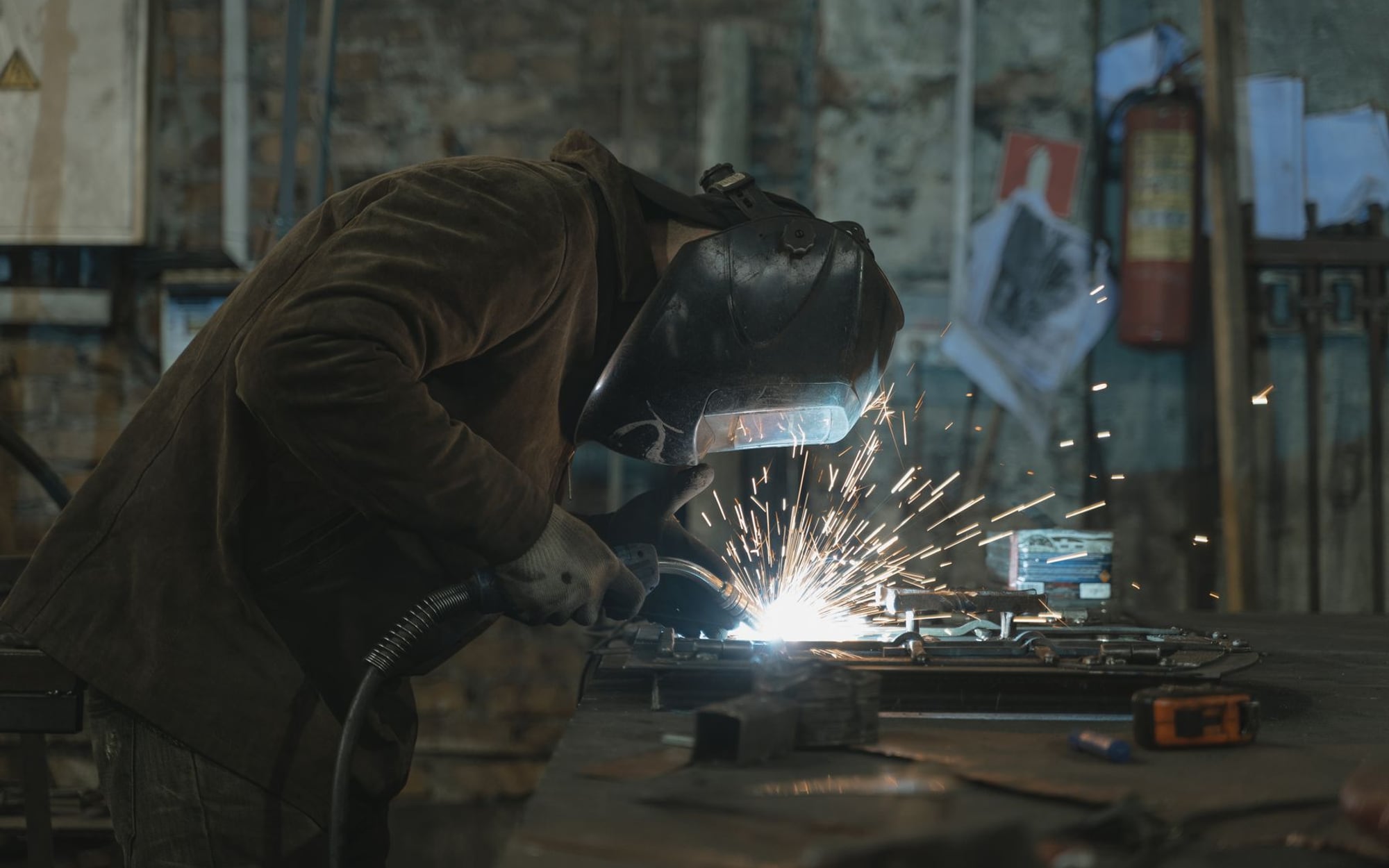 A welder in a full welding helmet working over steel in a dark shop, sparks scattering &mdash; representative imagery, not a specific Late Night Fabrication job